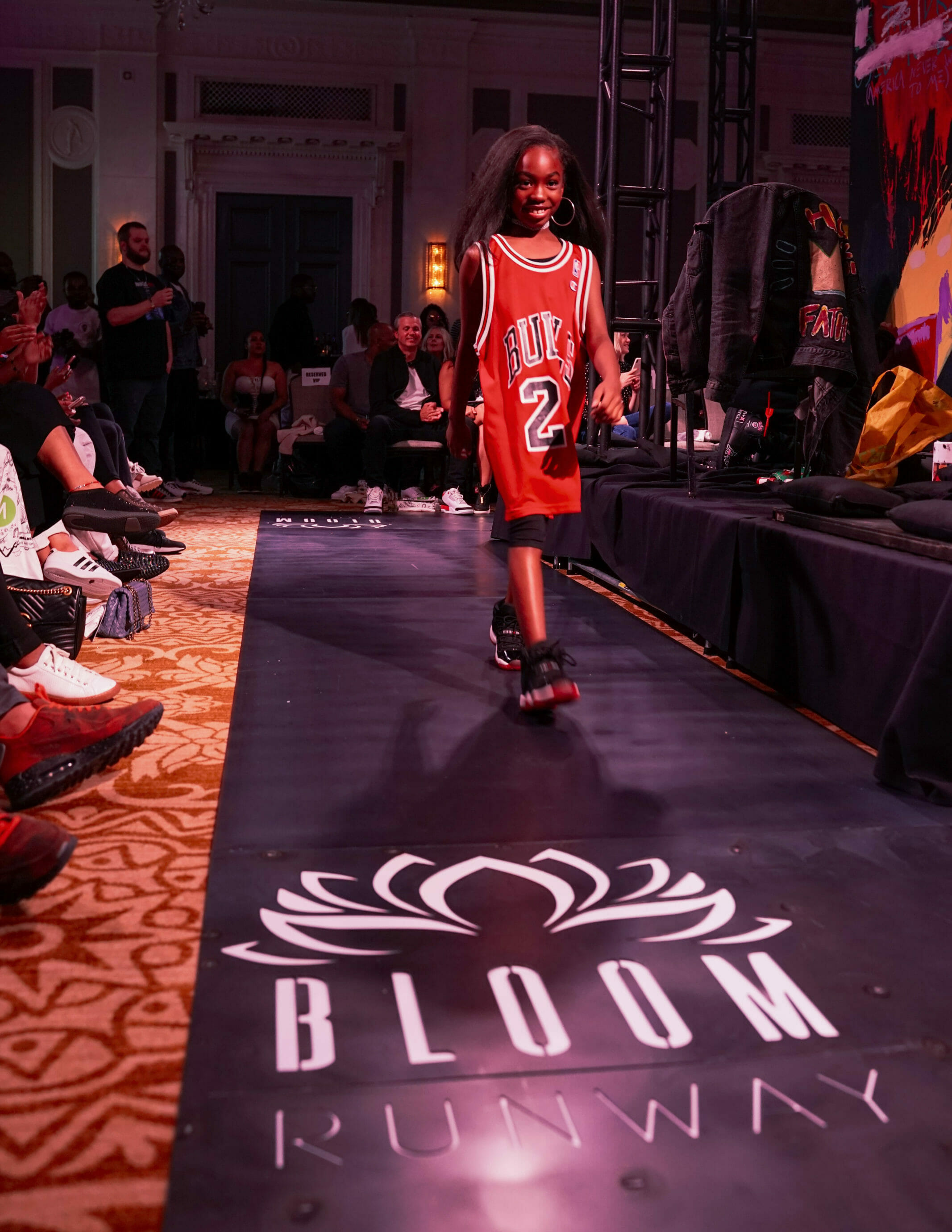 A child wearing a Bulls basketball jersey and gold hoop earrings walks confidently down a runway with “Bloom Beauty Collective” decals.