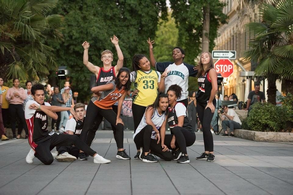 Teens pose excitedly in basketball jerseys and athletic wear in a public square with a crowd looking on and studio lights pointed at them.