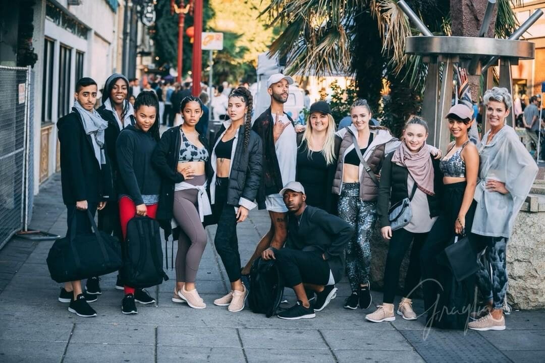 A group of 12 teens pose confidently in street wear in a busy public square.
