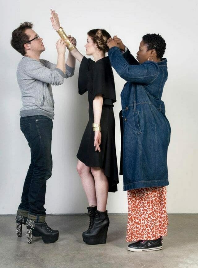 Abibat and another stylist work on a model, standing against a blank background. Abibat pins back the model’s hair, while the other stylist fixes a golden armband on them.