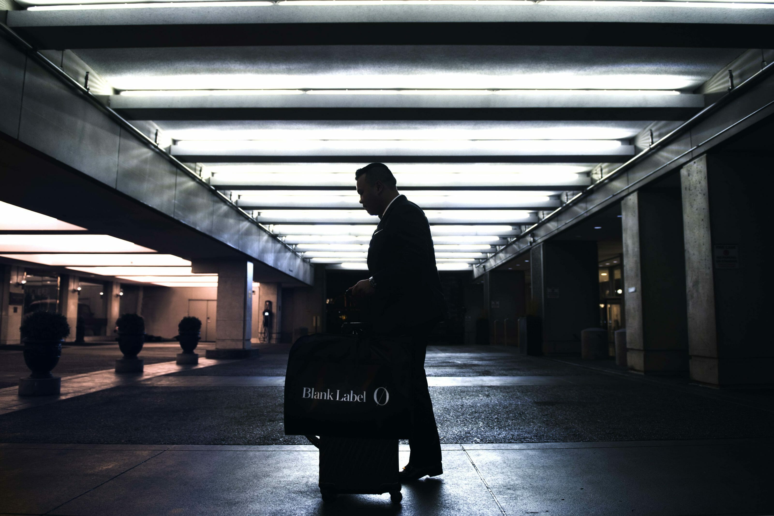 Man walking in a dimly lit garage, rolling a suitcase with a garment bag that reads “Blank Label.”