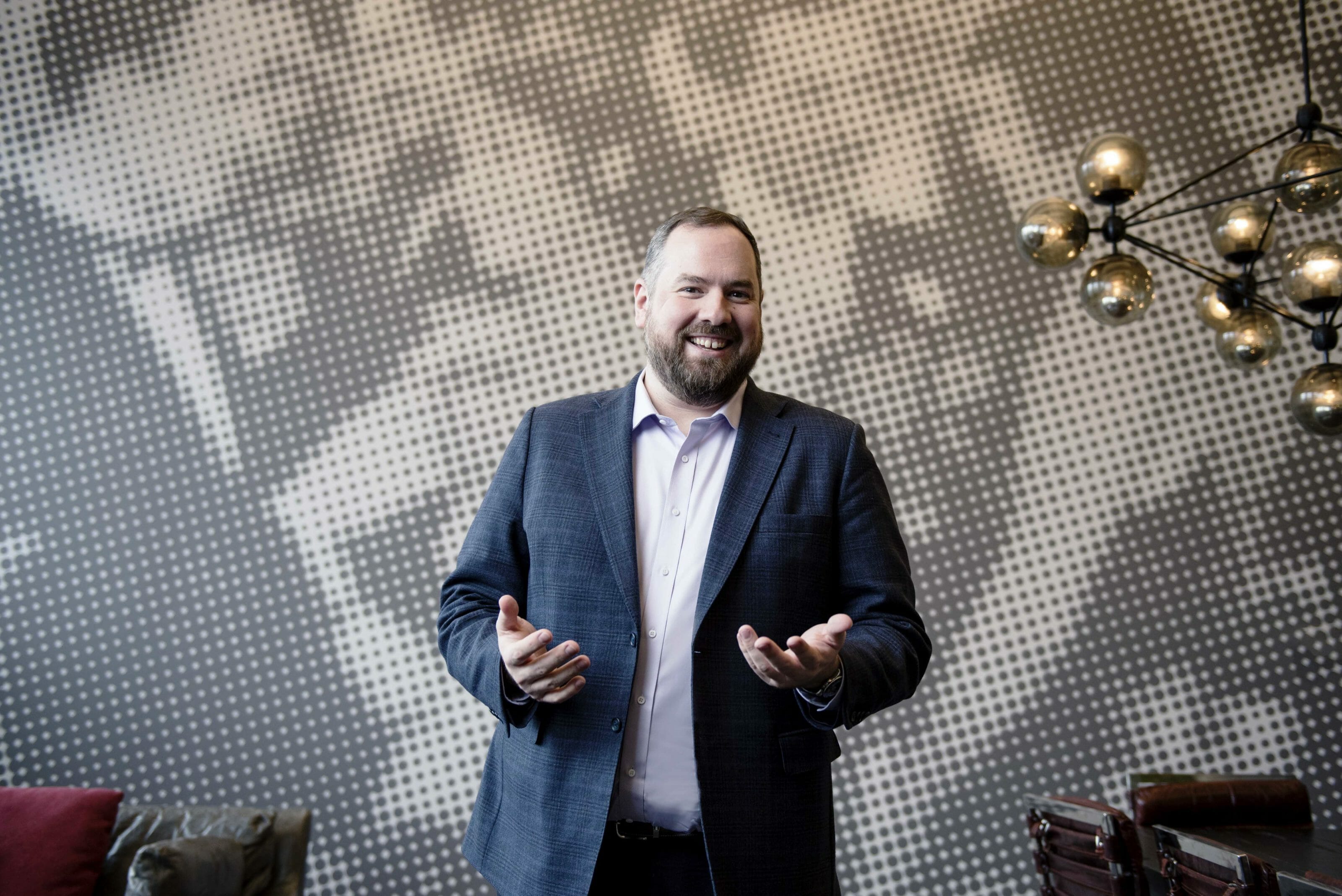 Man wearing an open plaid blazer with a lavender button-up shirt, smiling and posing in a contemporary styled room.