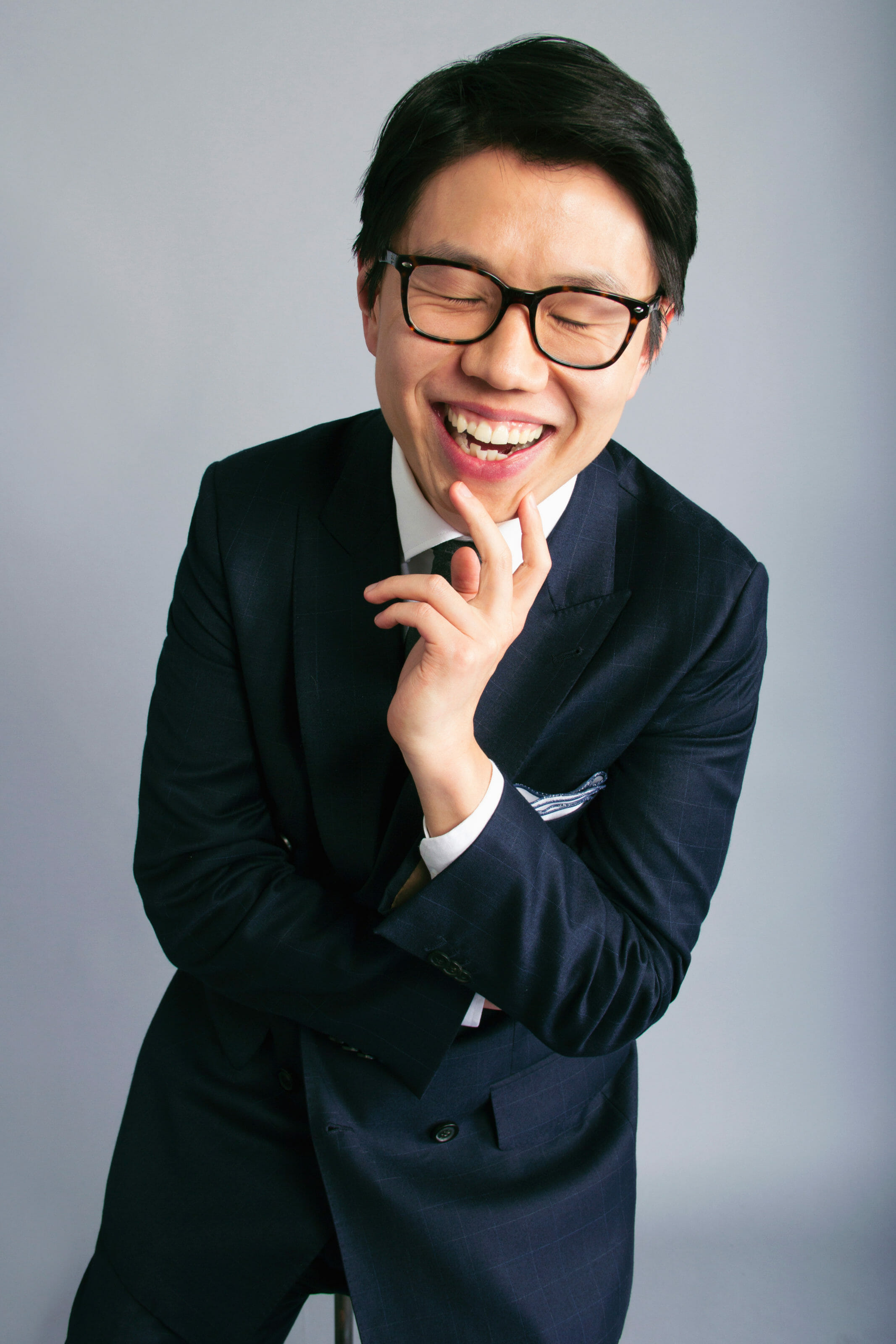 Fan Bi laughing, against a grey background wearing glasses and a well-tailored suit jacket.