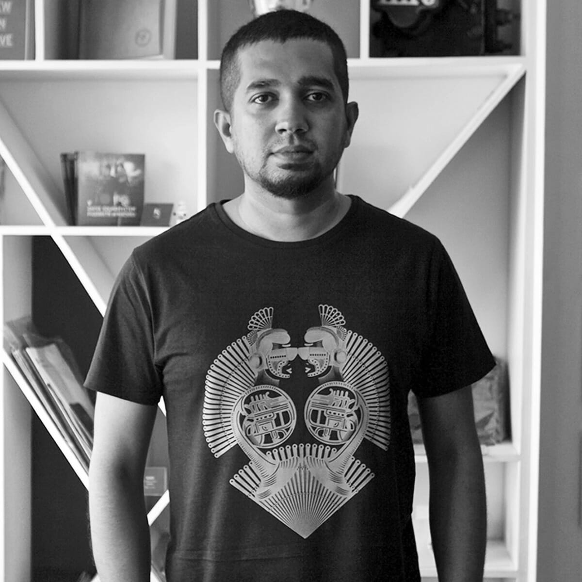 Black and white photo of Reshidev standing in front of a book shelf wearing a graphic t-shirt.