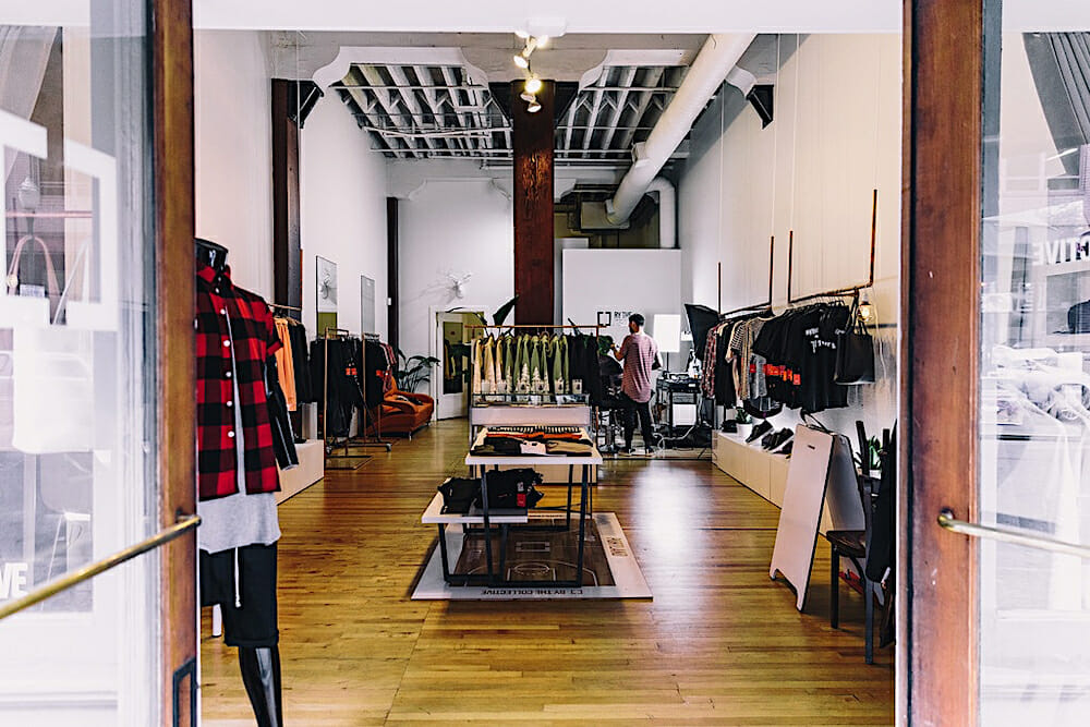 Wide shot photo of Jaefields retail space. In the shop there are wood floors with a table in the center and tall white walls holding racks for several hung t-shirts.