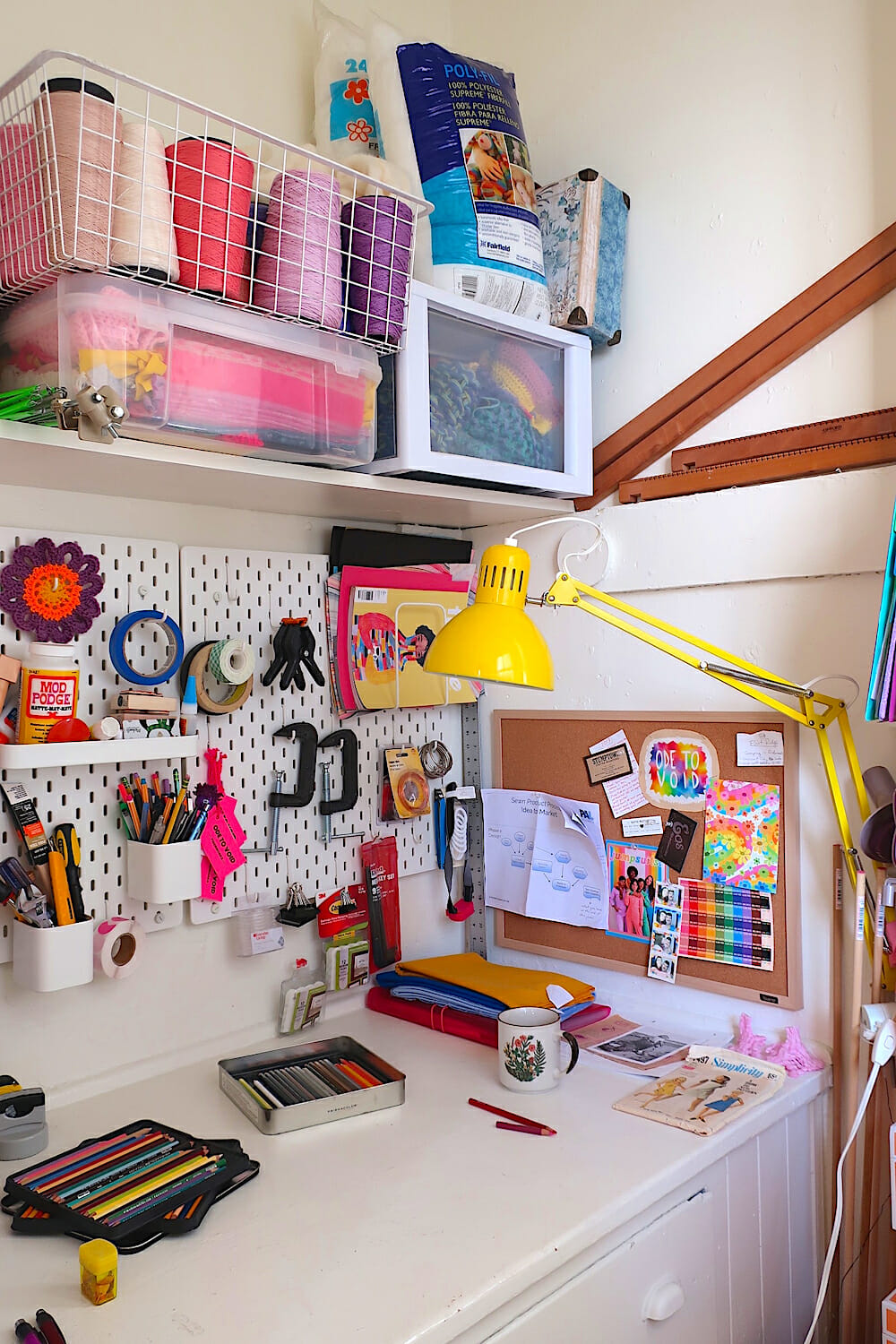 Photo of Kelsey's desk with a yellow lamp and colored pencils and a shelf holding pink spools of yarn.