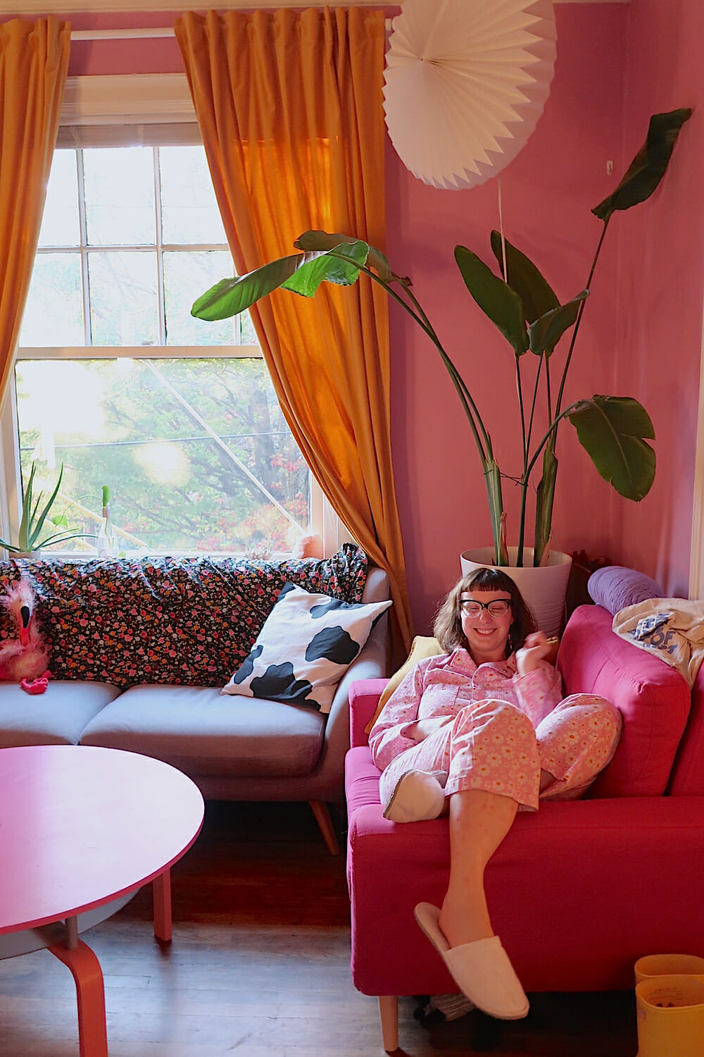 Photo of Kelsey smiling and laying back on a pink sofa in a pink living room with orange curtains.