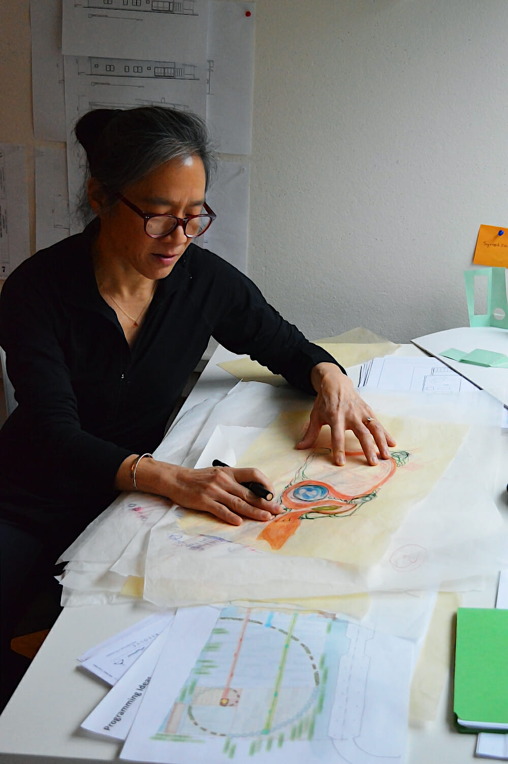 Suenn Ho Working Suenn leaning over a table with scattered papers, with a pen in her hand. Suenne is wearing a black outfit and reading glasses rest on her face.