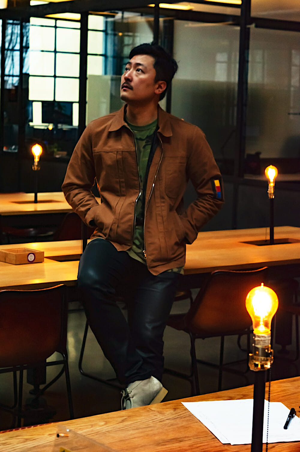 Portrait photo of Wookie leaning against a wooden table, hands in his pockets and right leg crossed over the left, while looking off to the distance. Around him are additional wooden tables with light bulbs sticking out. In the background are interior office glass windows. 