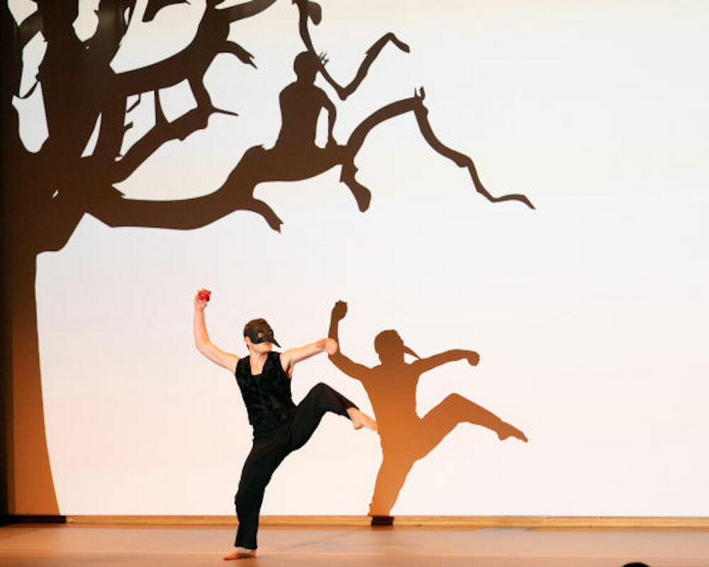 Male dancer in black mask poses with one leg raised against silhouette of a tree