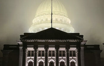 Stark columns of the Wisconsin State Capitol building are clear in the foreground while the lit-up dome behind is blurred by fog. 