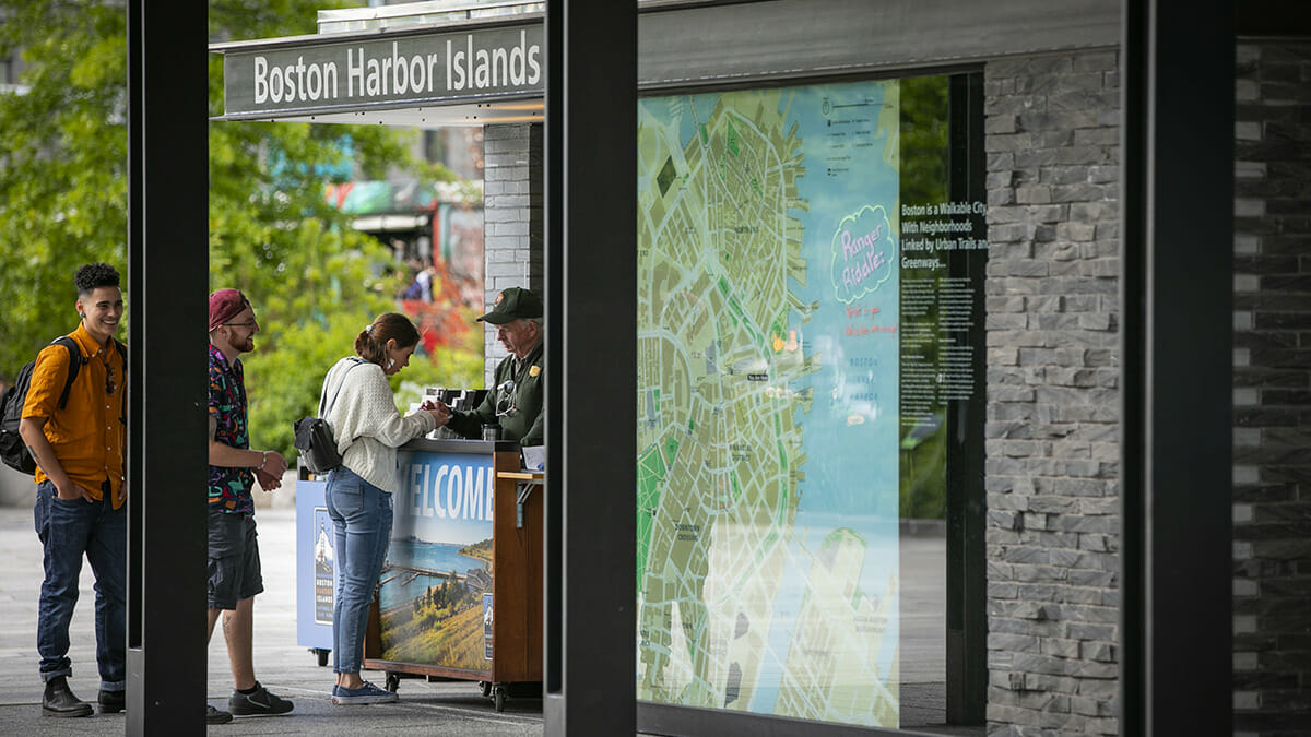 Ticket counter for Boston Harbor Islands with patrons waiting in line. Large scale map of the Boston waterfront to the left of the ticket counter. Rose Fitzgerald Kennedy Greenway Conservancy.