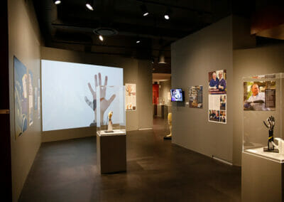 Exhibition installation showing various artifacts on display including the Complete Control System and a video projection of The Third Thumb in the background.
