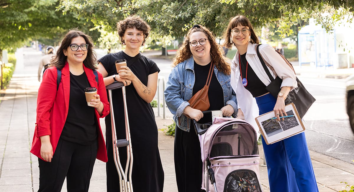Holst Architecture team: Four smiling women in black, blue, and red clothing standing on a wide sidewalk lined with green street trees. One drinks coffee, another is leaning on her crutches, one is pushing an orange cat (Fern) in a stroller, and the last holds a clipboard.