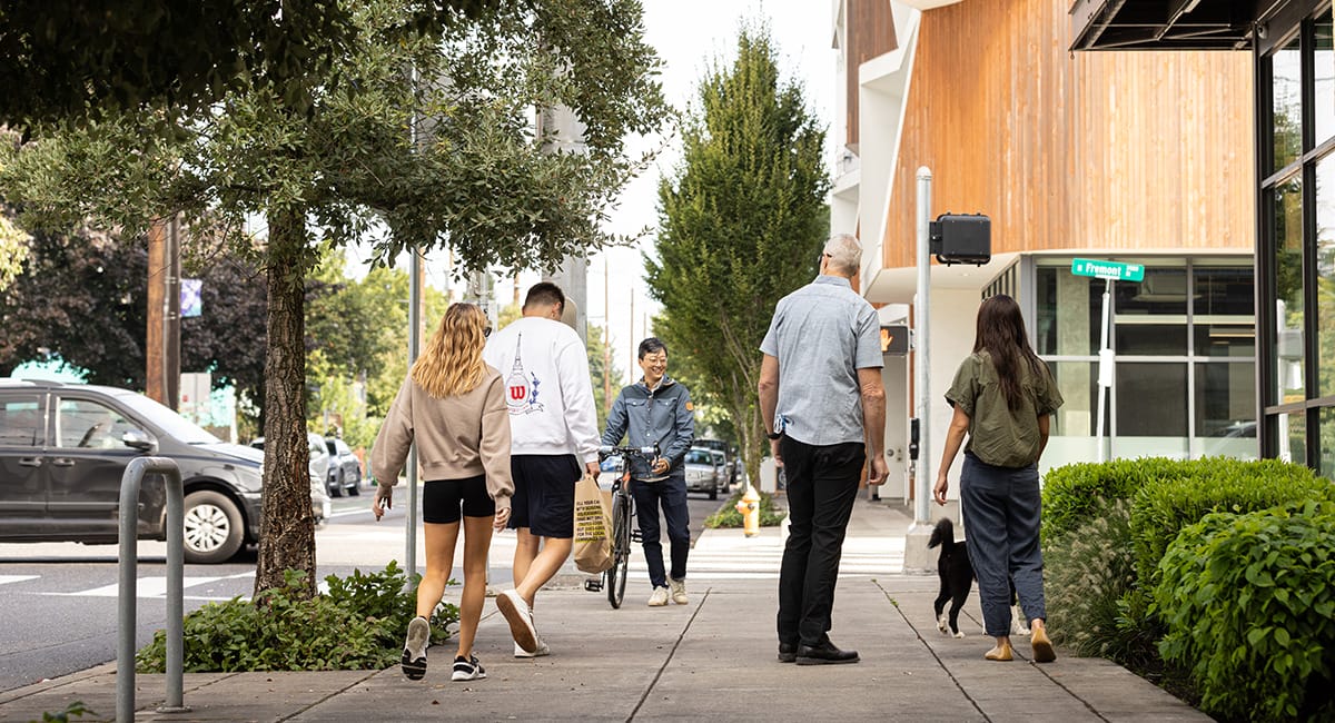 People pass each other on a wide sidewalk. One person rolls a bike, others carry groceries and walk a dog. Text points out "Extra-wide sidewalks help Deaf people, mobility device users, and plus size folks pass each other comfortably" and "Audible and tactile crosswalk signals help Blind and Deaf people cross the street."